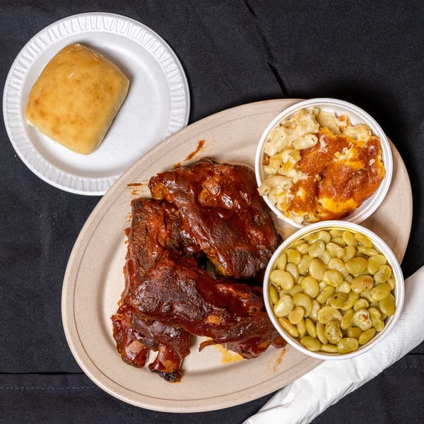A plate of BBQ ribs with mac and cheese, lima beans, and a dinner roll.