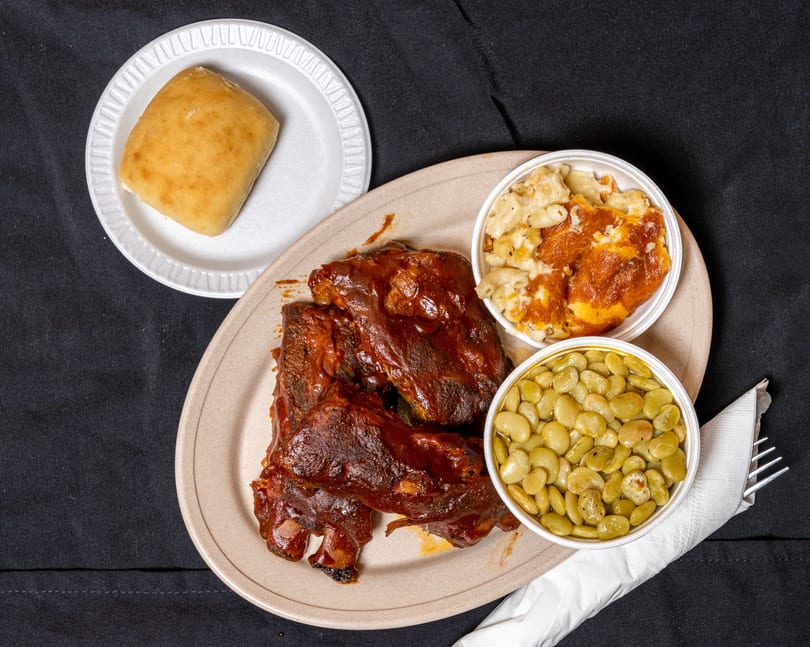 A plate of BBQ ribs with mac and cheese, lima beans, and a dinner roll.