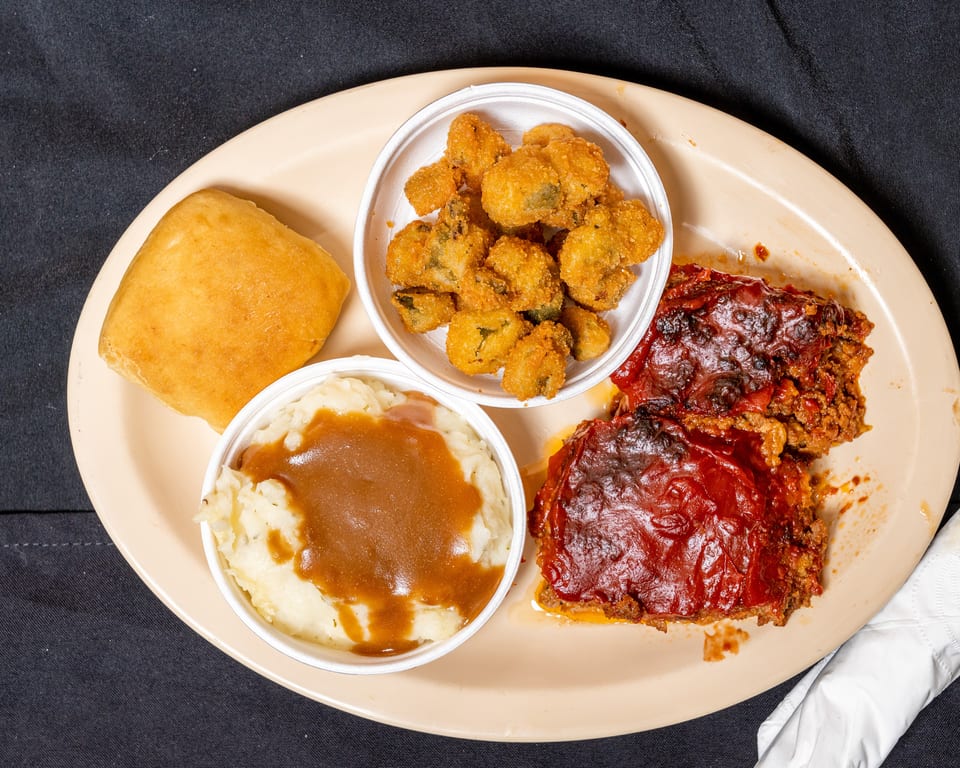 Meatloaf with red sauce, mashed potatoes with gravy, fried okra, and a dinner roll on a plate.
