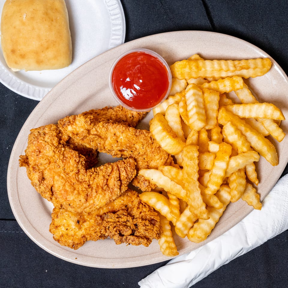 Crispy fried chicken tenders, crinkle-cut fries, ketchup, and a dinner roll on a dark surface.