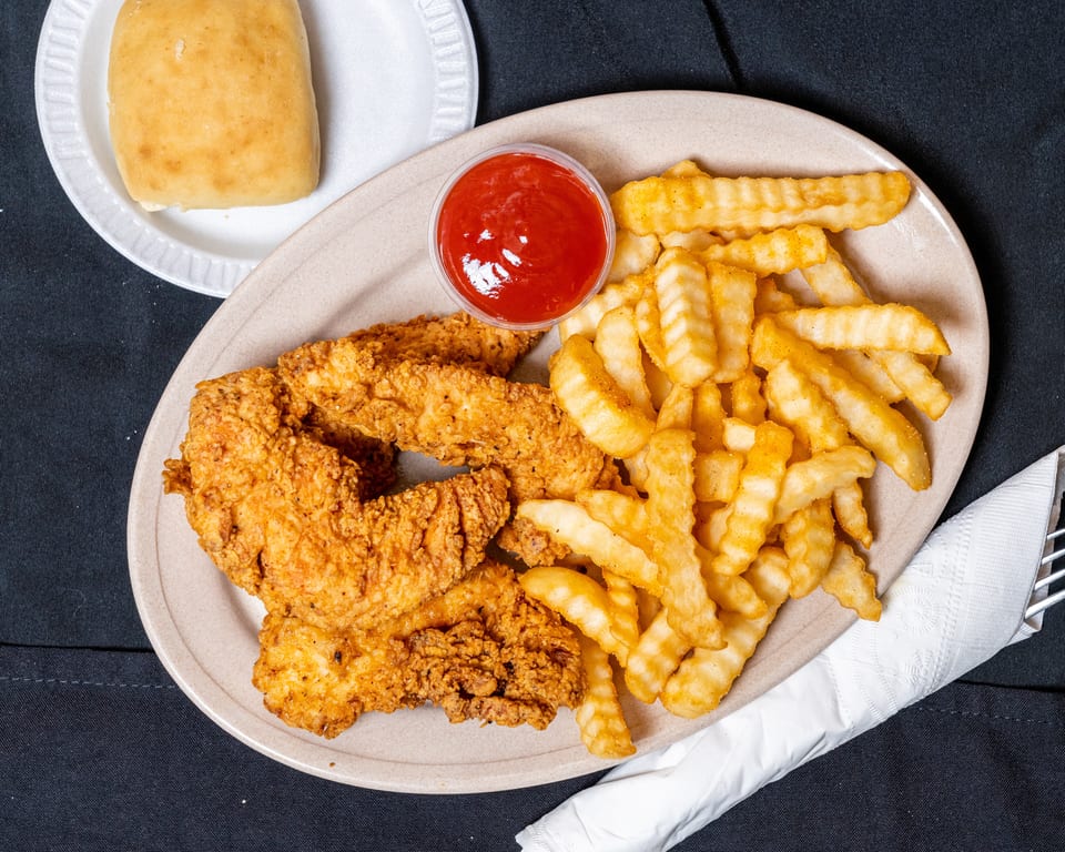 Crispy fried chicken tenders, crinkle-cut fries, ketchup, and a dinner roll on a dark surface.