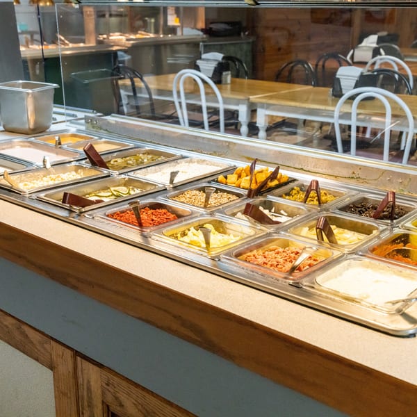 A salad bar with various toppings in metal containers, with dining tables and chairs in the background.