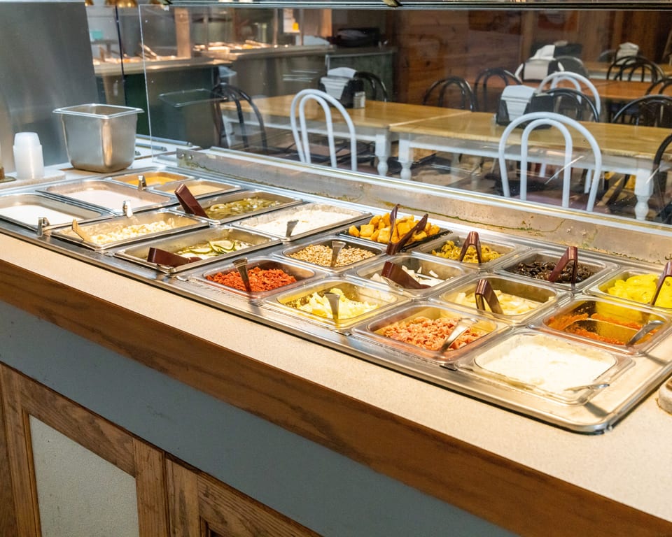 A salad bar with various toppings in metal containers, with dining tables and chairs in the background.