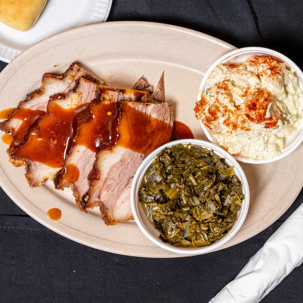 Sliced pork with barbecue sauce, collard greens, and potato salad on a plate.
