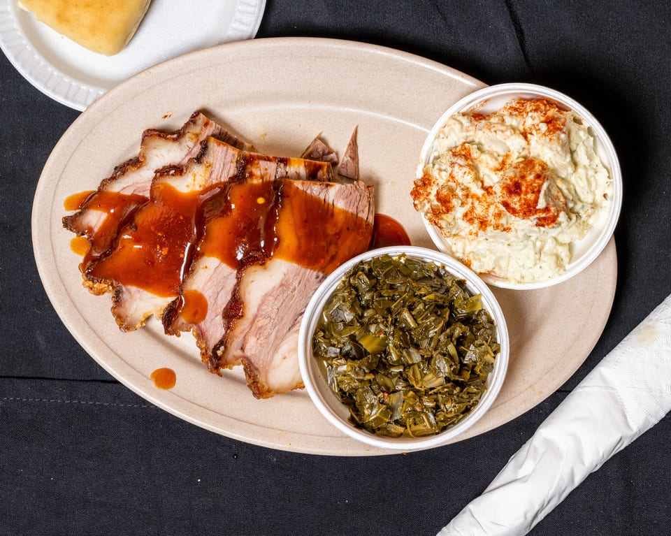 Sliced pork with barbecue sauce, collard greens, and potato salad on a plate.